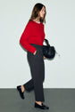 A woman with long brown hair wears a red sweater, gray pantalon droit à coupe droite, and black loafers. She carries a black textured handbag and walks against a plain light background.