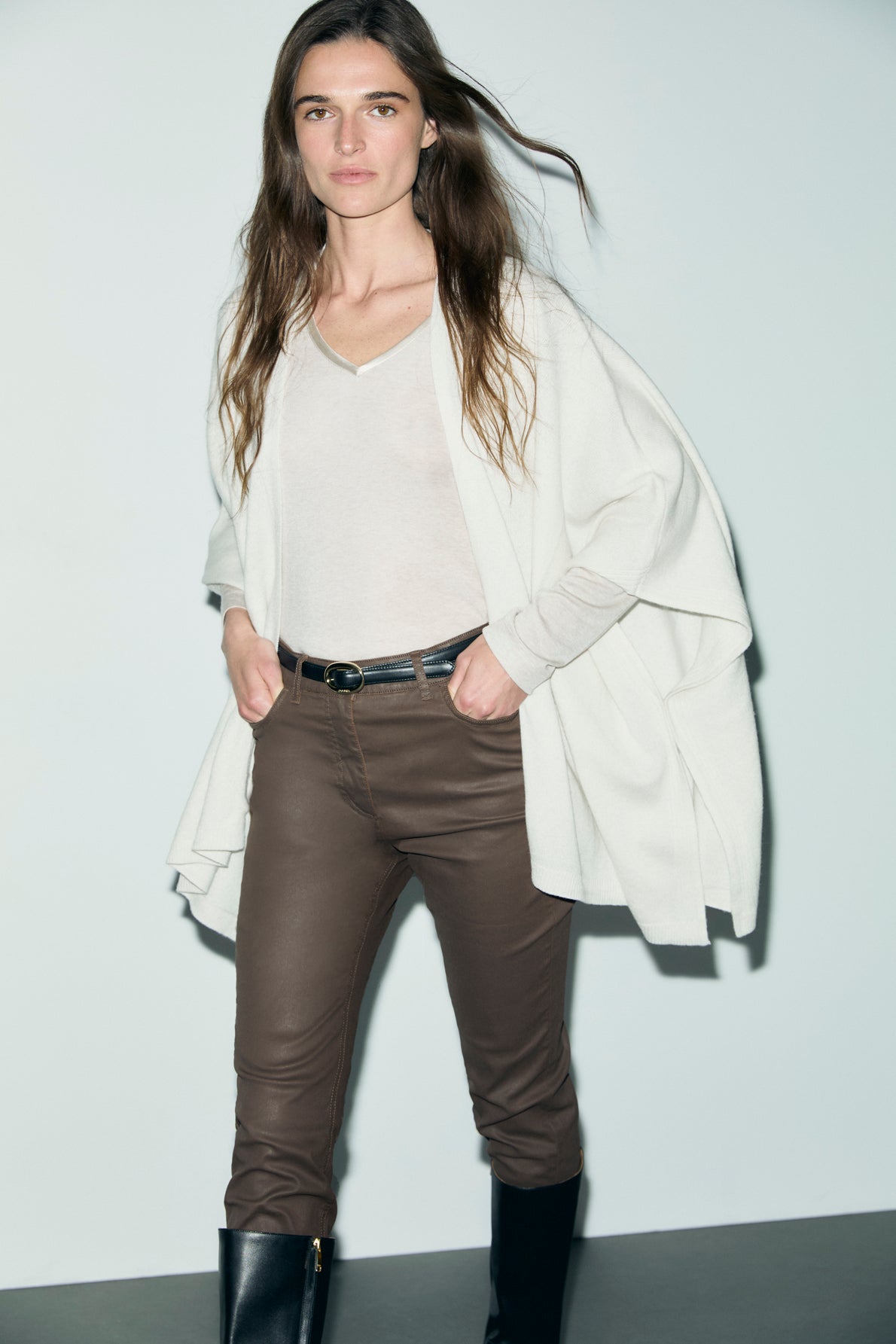 Woman with long brown hair wearing a cream sweater, light top, brown pants effet cuir, and black boots stands against a plain light background, looking at the camera.