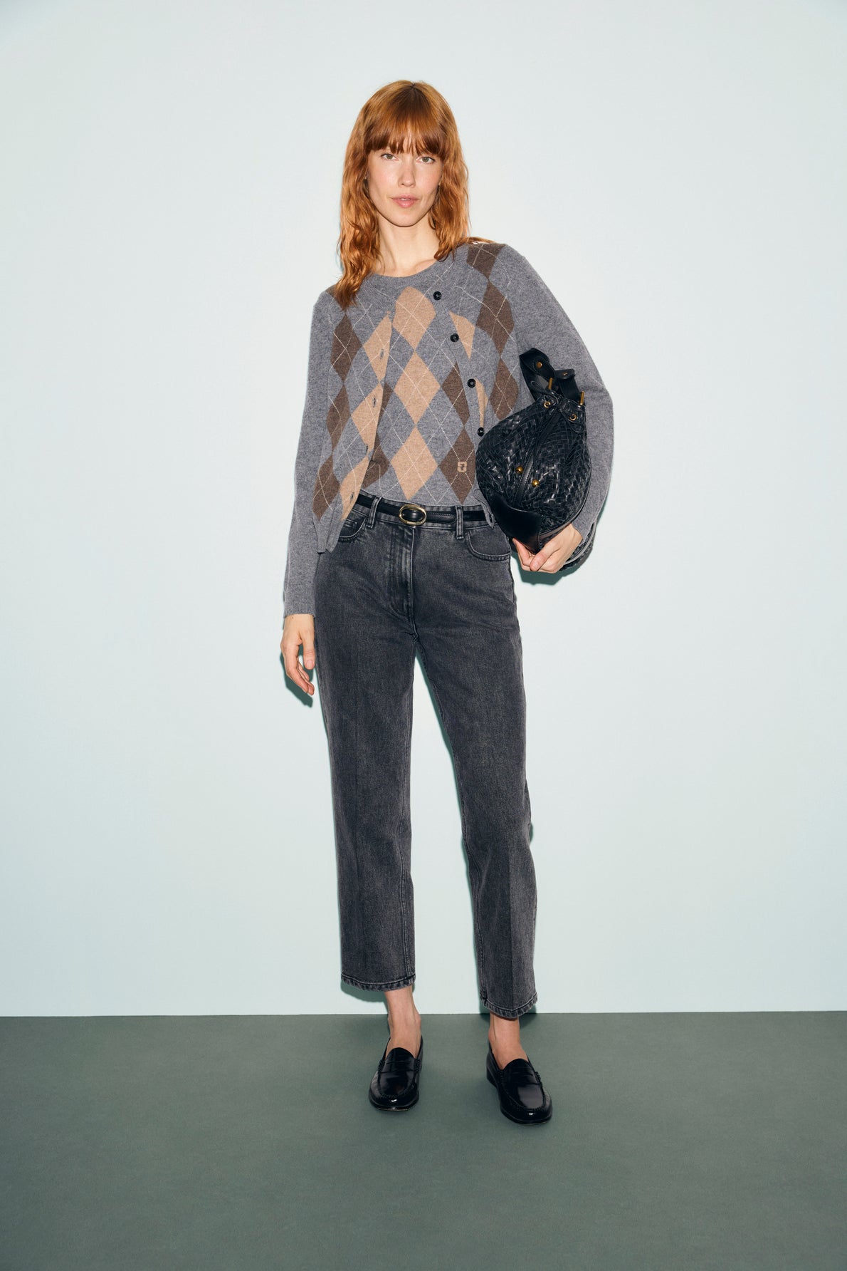 A woman with red hair wears a gray argyle sweater, denim anthracite jean droit modèle cinq poches, and black loafers, holding a black textured handbag, standing against a plain light blue background.