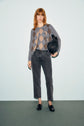 A woman with red hair wears a gray argyle sweater, denim anthracite jean droit modèle cinq poches, and black loafers, holding a black textured handbag, standing against a plain light blue background.