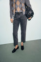 Person wearing a gray argyle sweater, denim anthracite jean droit modèle cinq poches, black loafers, and holding a black woven handbag, standing against a pale background. The person’s head is not visible in the image.