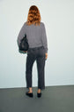 A person with long reddish hair, wearing a gray sweater and denim anthracite jean droit, stands facing a light wall. They hold a black textured handbag and wear black shoes.