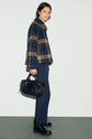 A person wearing a blue and tan plaid jacket, a pantalon slim bleu marine, and black boots stands holding a black handbag against a plain white background.