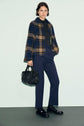 Woman stands against a light background wearing a blue and brown plaid jacket, dark turtleneck, and a pantalon slim bleu marine for a sophisticated silhouette. She completes the look with black shoes, holds a black handbag, and has short, wavy brown hair.