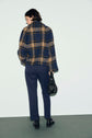 Person standing against a plain background, wearing a blue and tan plaid jacket paired with a pantalon slim bleu marine, black shoes, and holding a small black handbag. The person is facing away from the camera.
