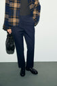 Person wearing a pantalon slim bleu marine, a blue and brown plaid jacket, and black shoes, holding a black handbag, standing against a plain light wall. The person’s face is not visible.