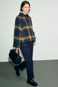 A woman wearing a blue and tan plaid jacket, a pantalon slim bleu marine, and black shoes walks while carrying a black handbag, set against a plain light background.