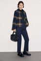 A person stands against a plain background wearing a blue and brown plaid jacket, dark top, pantalon slim bleu marine, black shoes, and holding a black handbag.