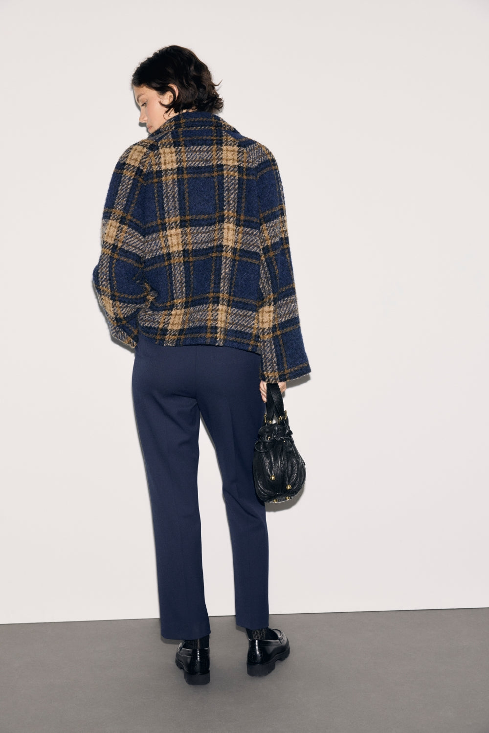 A person stands facing away, wearing a blue and tan plaid jacket, a pantalon slim bleu marine, and black shoes, holding a small black bag. The background is plain and light-colored.
