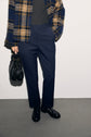 A person wearing a pantalon tailleur femme—specifically, a pantalon slim bleu marine—paired with a grey top, a blue and tan plaid jacket, and black shoes holds a black handbag, standing against a plain light background.