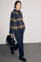 A woman wearing a blue and beige plaid jacket, a pantalon slim bleu marine, and black shoes walks while carrying a black handbag against a plain light background.
