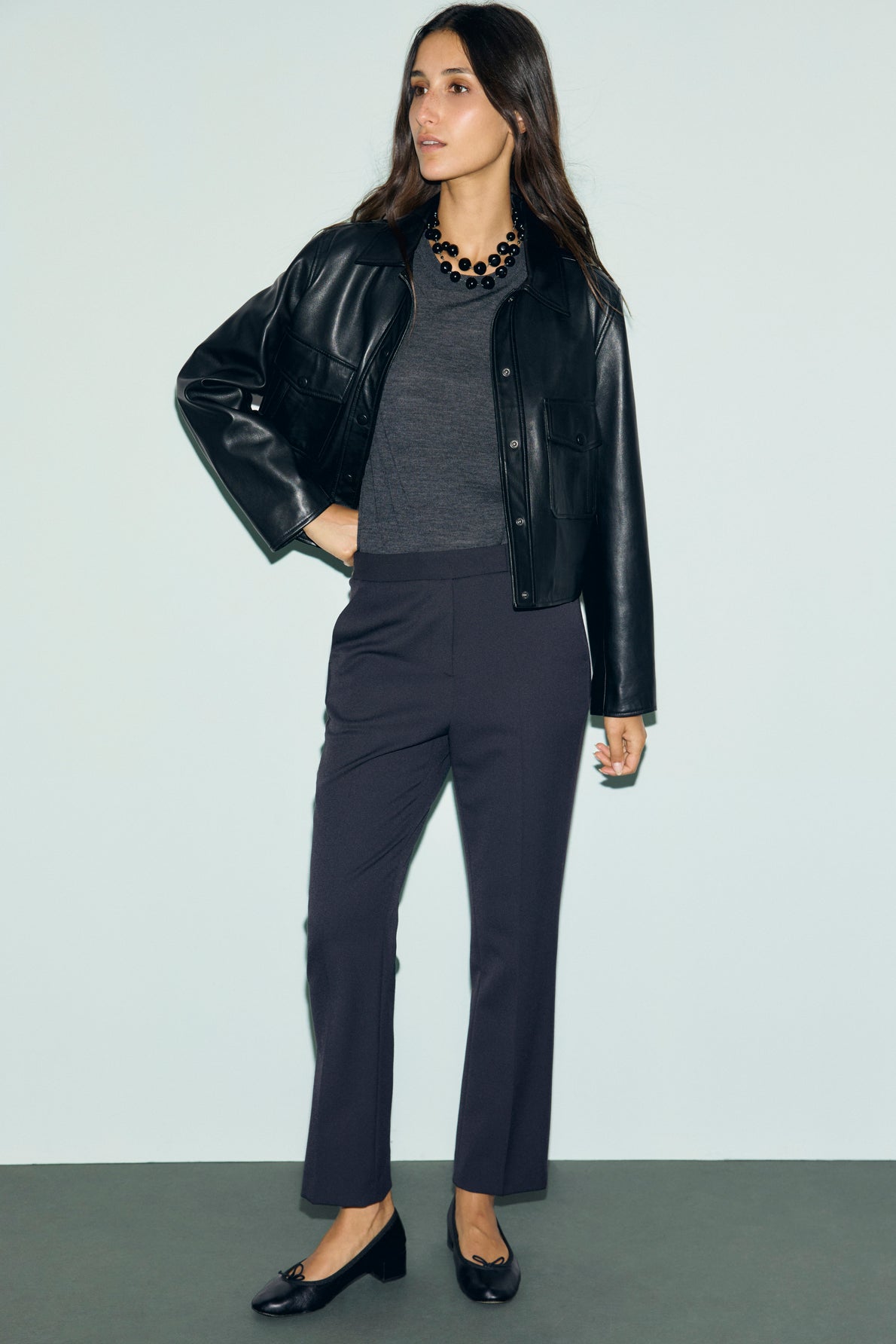 Woman standing against a plain background, wearing a black leather jacket, dark gray top, and a pantalon de tailleur coupe droite in velours bleu nuit. She pairs the look with black flats and a black beaded necklace. She has long brown hair and a neutral expression.