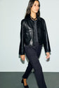 A woman with long dark hair walks indoors wearing a black leather jacket, a coupe droite pantalon de tailleur in velours bleu nuit, black shoes, and a chunky black necklace, against a plain light-colored wall.