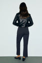 A person with long brown hair, wearing a black leather jacket, a pair of velours bleu nuit pantalon de tailleur à coupe droite, and black shoes, stands facing a light-colored wall with their back to the camera.