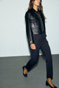 A woman walks indoors wearing a black leather jacket, a coupe droite pantalon de tailleur, and black low-heeled shoes, with her long hair loose and face partially out of view.