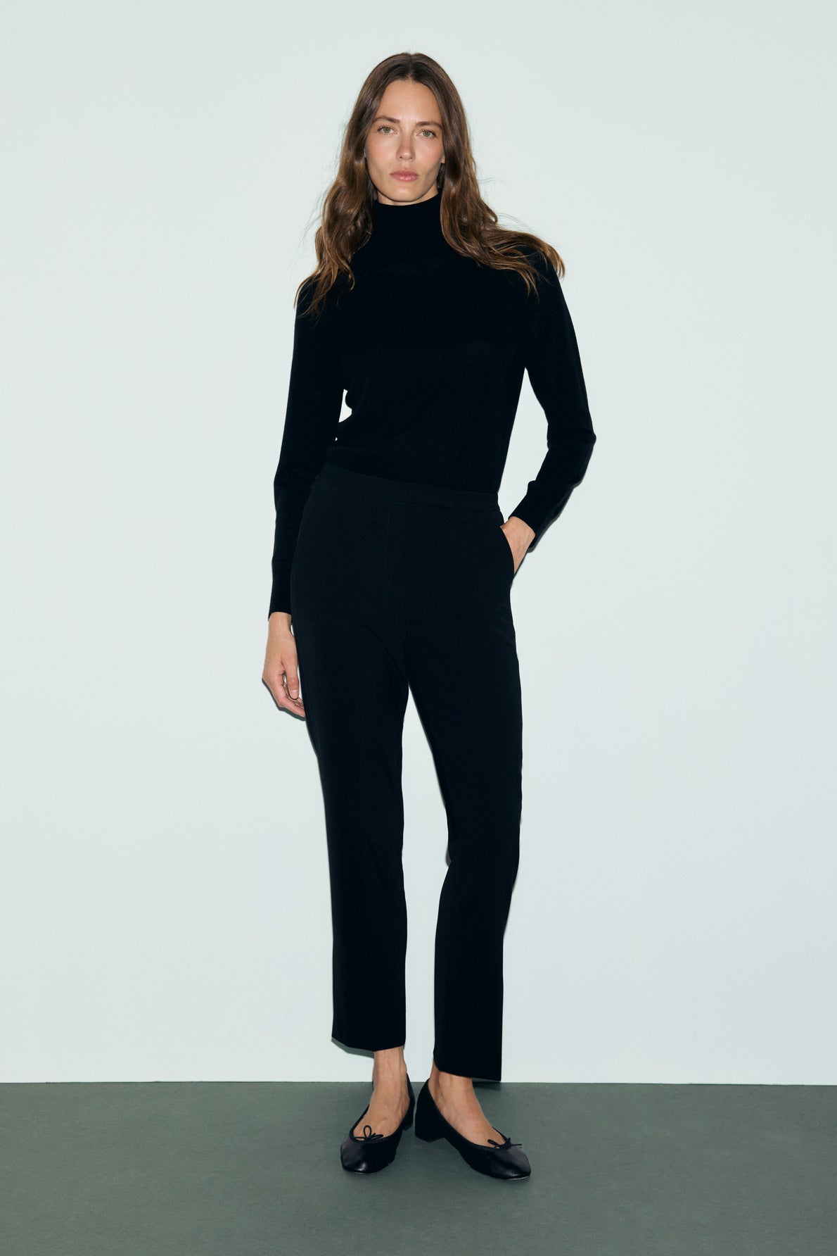 A woman stands against a plain white background, wearing a black turtleneck sweater, a pantalon de tailleur coupe slim longueur 7/8, and black shoes. She has long brown hair and a neutral expression, with one hand in her pocket.