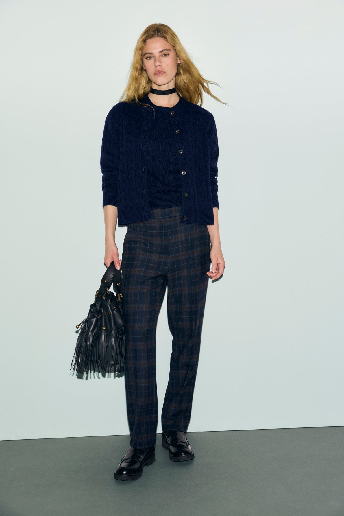 A woman with long blonde hair wears a navy cardigan, black top, and a pantalon de tailleur coupe droite bleu marine à carreaux, paired with black loafers and holding a black fringed handbag. She stands against a plain light background.