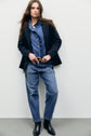 Woman stands against a plain wall wearing a navy blazer featuring une grande poche plaquée, a blue denim shirt with ruffle detail, coupe ample blue jeans in the jean barrel style, a black belt, and black ankle boots. She has long brown hair and a neutral expression.