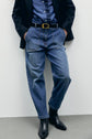 Person wearing blue denim jeans with a coupe ample fit, a blue denim shirt featuring a grande poche plaquée, a black blazer, a black belt with a gold buckle, and black boots, standing against a light-colored wall with hands in pockets.