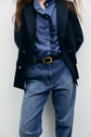 Person wearing a navy blazer over a ruffled blue denim shirt, paired with coupe ample high-waisted jeans and a belt, standing against a plain light background. Their face is not visible.