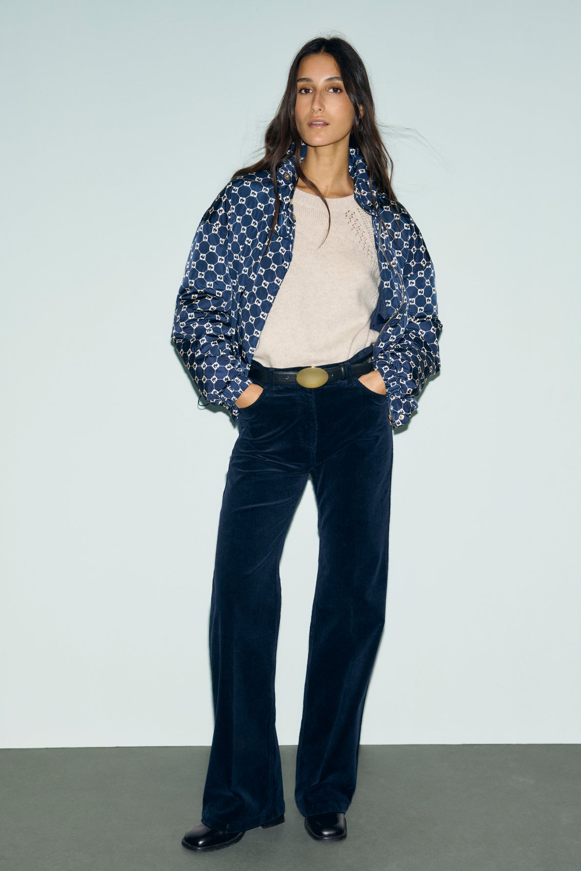 A woman stands against a light blue wall, wearing a blue patterned puffer jacket, beige shirt, and bleu marine pantalon large en velours côtelé with a belt. Her hands are in her pockets and she finishes the look with black shoes.