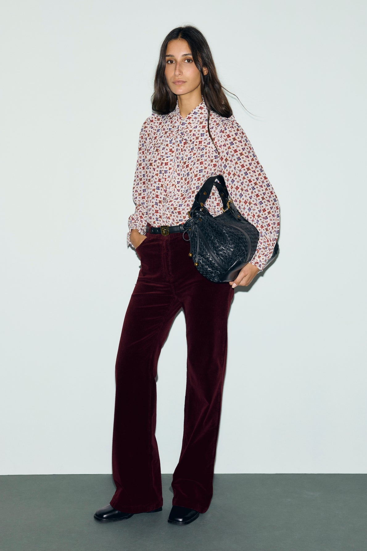 Woman standing against a plain wall, wearing a floral blouse, bordeaux pantalon velours côtelé large and black shoes, holding a large black textured bag in her left hand.