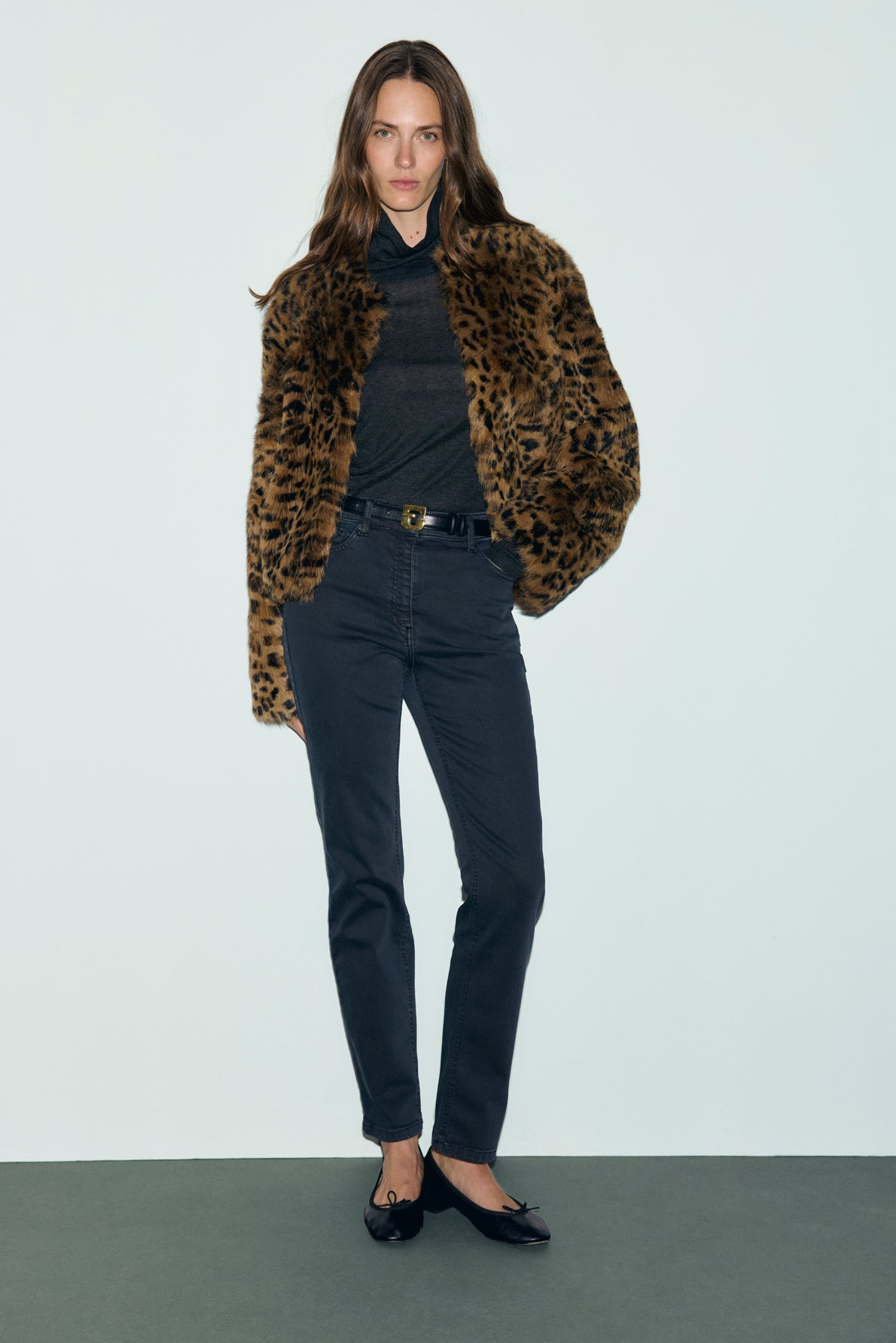 A woman stands against a plain background wearing a leopard print faux fur jacket, sheer black top, and a pair of jean slim noir with a belt. Her black flats complete the look, while her hair falls loosely as she casually slips her hands into her pockets.