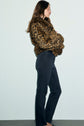 A woman stands in profile against a plain background, wearing a leopard print faux fur jacket and a pair of jean slim noir, with her arms folded and her long brown hair loose.