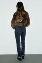 A person with long brown hair, wearing a leopard print jacket, black jean slim noir with a coupe slim fit, and black shoes, stands facing away from the camera against a plain background.