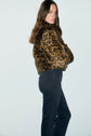A person with long brown hair wears a leopard-print faux fur jacket and black pants in a coupe slim fit, standing sideways against a plain light background, looking over their shoulder.