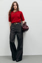 A woman in a red short-sleeve sweater, wide-leg jean évasé trousers, and black shoes stands against a plain wall, holding a large burgundy bag.