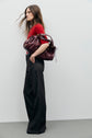 A woman in wide-leg jean évasé dark trousers and a red top stands against a plain light wall, holding a large burgundy handbag over her shoulder with one hand in her pocket.
