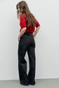 A person with long brown hair wears a red short-sleeve knit top and high-waisted black jean évasé pants, standing sideways with hands in pockets against a plain light background.