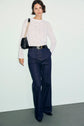 Woman standing against a plain background, wearing a white long-sleeve lace top, dark high-waisted jean évasé, a black belt, black shoes, and a black shoulder bag with a chain strap.