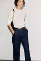 A person wearing a white textured long-sleeve top, dark blue jean évasé high-waisted jeans, and a black belt, with a black shoulder bag featuring a gold chain strap, stands against a plain light background.