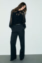 Woman standing against a plain wall wearing a sheer black blouse and a pantalon de tailleur coupe droite with large wide-leg trousers, hands in pockets, looking down with long hair over her shoulders.