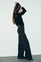 A person with long hair, wearing a black lace blouse and a pantalon de tailleur coupe droite, stands sideways with arms raised above their head against a plain background.