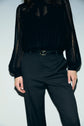 Person wearing black pleated high-waisted pantalon de tailleur with a coupe droite and a sheer black lace blouse with long sleeves, standing against a plain light background. Only the torso and legs are visible.
