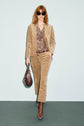 A woman with red hair wears sunglasses, a tan corduroy suit featuring a pantalon velours beige, a paisley blouse, brown boots, and carries a brown handbag, walking confidently against a light blue background.