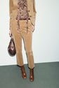Person wearing a pantalon velours beige styled as a tan corduroy suit with a paisley top, brown belt, brown ankle boots, and holding a dark brown handbag. The photo is cropped at the shoulders.