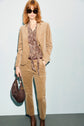 A woman in sunglasses wears a tan corduroy suit with a paisley blouse, brown boots, and holds a dark brown handbag, standing against a light background. Her look is completed by a chic pantalon velours beige, adding effortless elegance to her outfit.