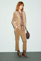 A person with wavy, reddish-brown hair wears a pantalon velours beige corduroy suit, a patterned blouse, brown ankle boots, and carries a brown handbag, standing against a pale blue background.