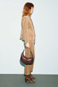 A woman in a tan corduroy suit featuring a pantalon velours beige stands sideways, holding a dark brown handbag and wearing brown heeled boots, against a plain background.
