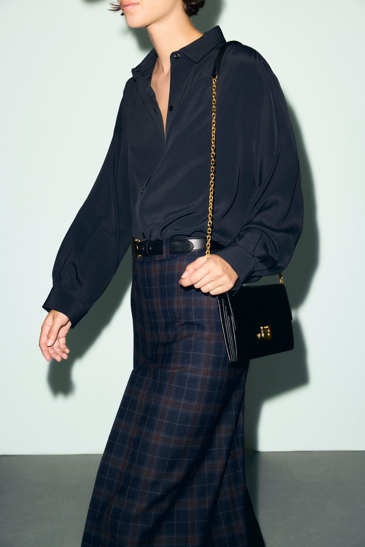 A person wearing a black blouse and a style preppy bermuda jupe à carreaux, carrying a black purse with a gold chain, walks against a light background.