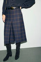 A person wears a dark plaid knee-length skirt reminiscent of a bermuda jupe à carreaux, paired with a black long-sleeve shirt, a black belt, and black knee-high boots, effortlessly embodying a style preppy aesthetic while standing against a neutral background.