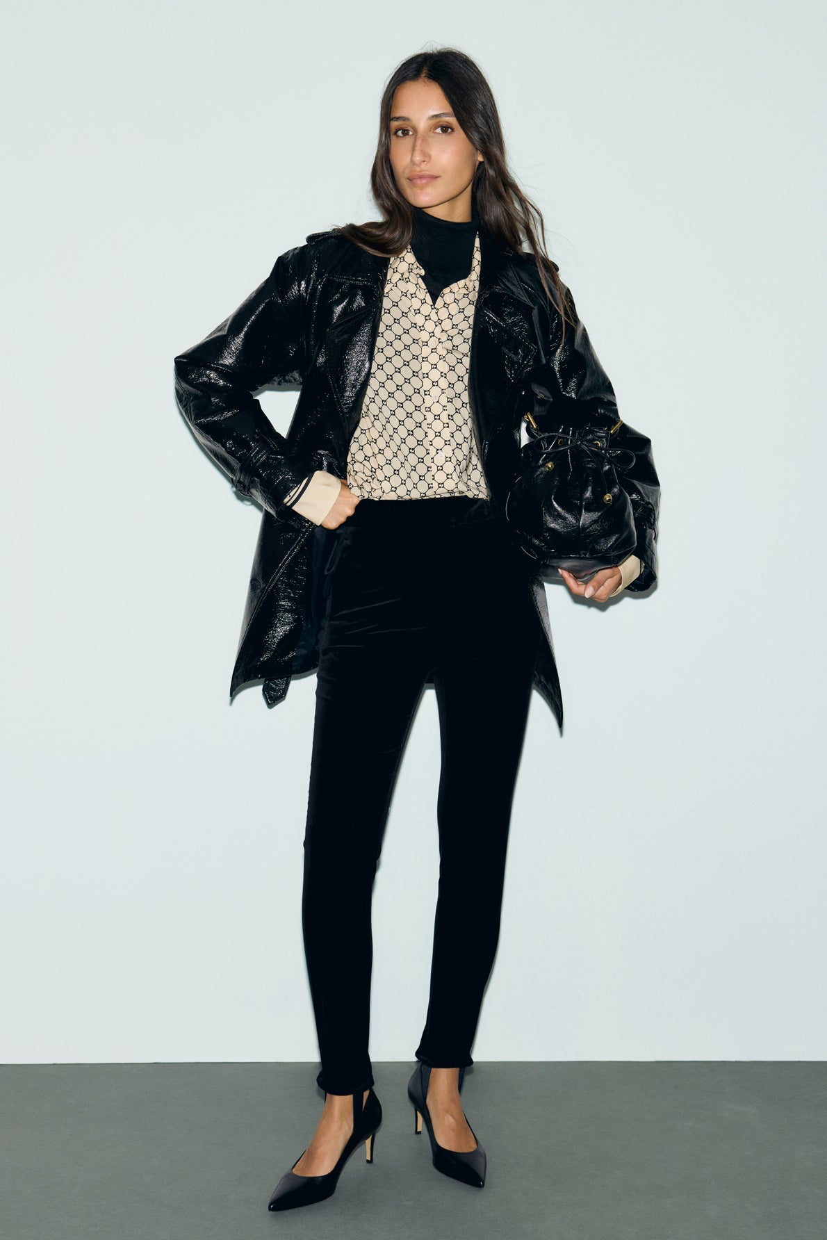A woman stands against a plain background wearing a black shiny coat, patterned sweater, black pants, pointed high heels, and holding a black handbag.