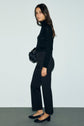 A woman stands sideways against a plain wall, dressed in a black turtleneck and a chic pantalon tailleur noir 7/8, paired with matching black shoes. She holds a small black bag and has long brown hair.