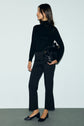 A woman stands against a plain background wearing a black long-sleeve top, a sleek pantalon tailleur noir, and black flats, holding a black bag under her arm.