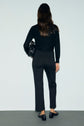 A woman with long brown hair stands facing a plain wall, wearing a black sweater, a pantalon tailleur noir 7/8, and black loafers, holding a black bag under her arm.