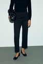 A person wearing a black long-sleeve top, a pantalon tailleur noir 7/8, and black flats stands on a grey floor, holding a black handbag. Their head is not visible in the image.